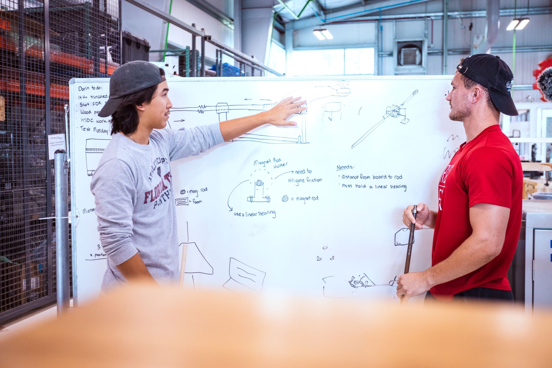 Two students discussing a project in front of a whiteboard filled with engineering drawings and notes in a workshop setting